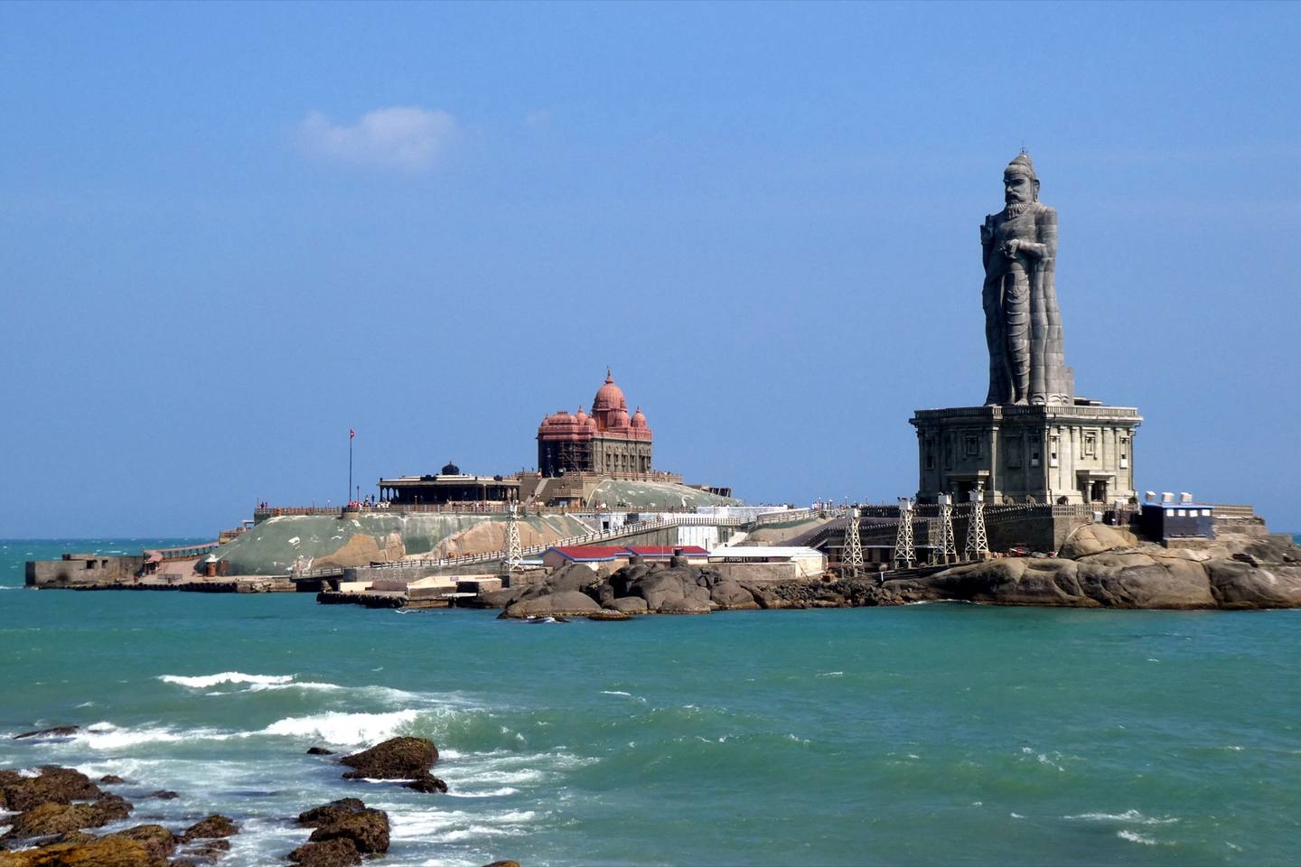 The Vivekananda Rock Memorial and the Thiruvalluvar Statue on their rocks in the sea off Kanyakumari, with waves breaking on the shore rocks under a clear blue sky