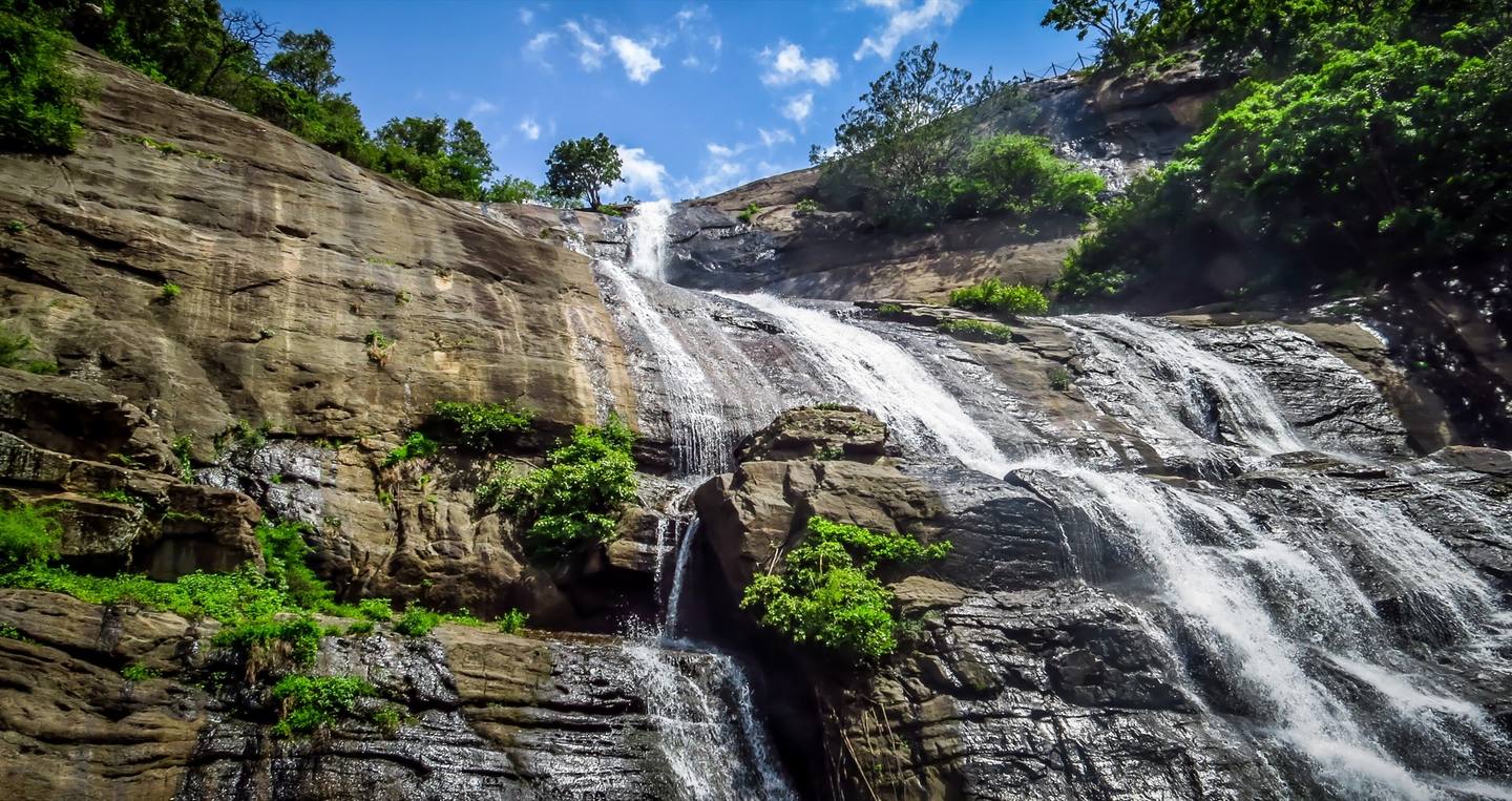Water from the Courtallam falls pouring down a wide rock face, with green trees along the top and a blue sky with small clouds